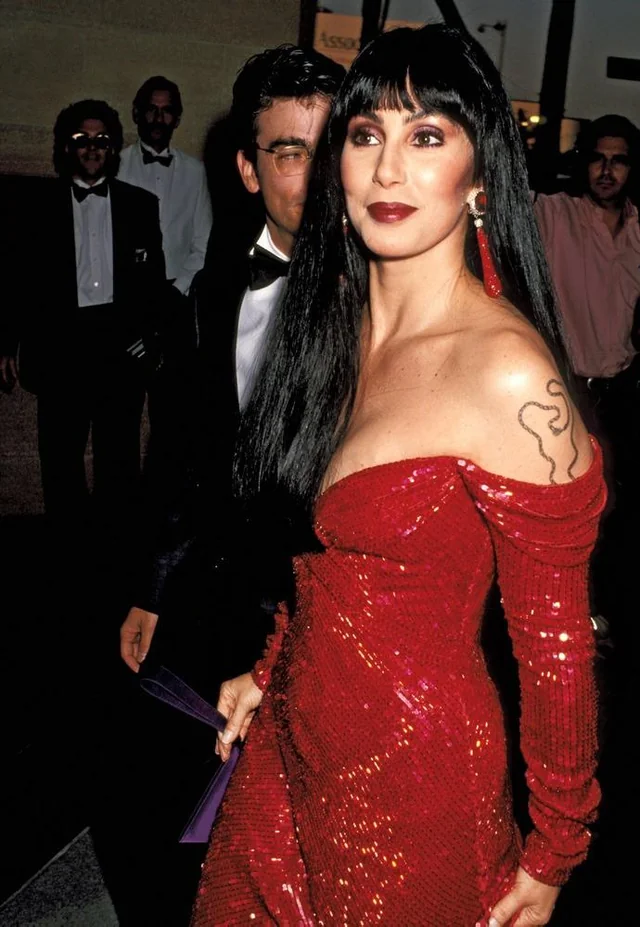 A woman with long black hair, wearing a sparkling off-the-shoulder red dress and red earrings, stands confidently at an event. Several people in formal attire are visible in the background.