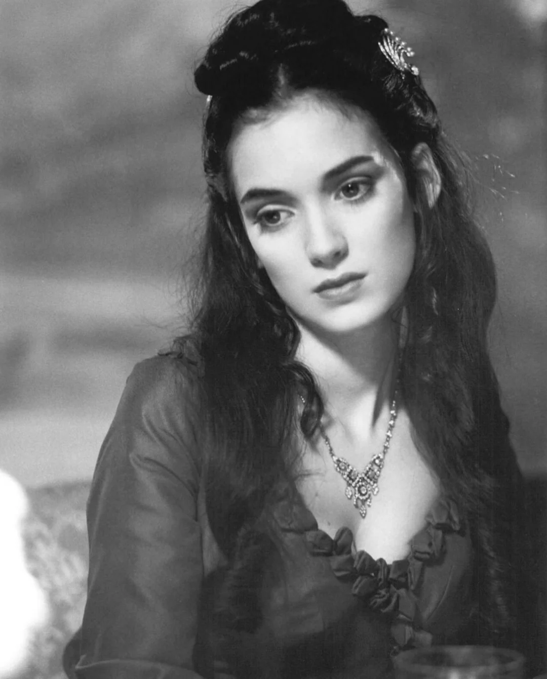 A young woman with long dark hair, dressed in a ruffled, old-fashioned gown, gazes thoughtfully to the side. She wears a decorative necklace and a hair clip, with a soft, blurred background behind her. The image is in black and white.