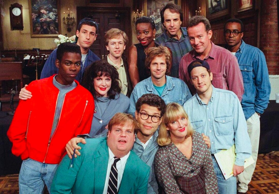 A group of twelve people pose together indoors, smiling at the camera. They are standing in rows inside a large room with wooden walls and paintings visible in the background.