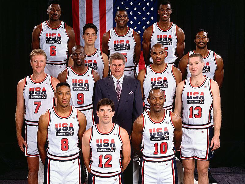 A basketball team in USA uniforms poses in front of American flags with their coach in the center. The team members are standing and kneeling, smiling for a group photo.
