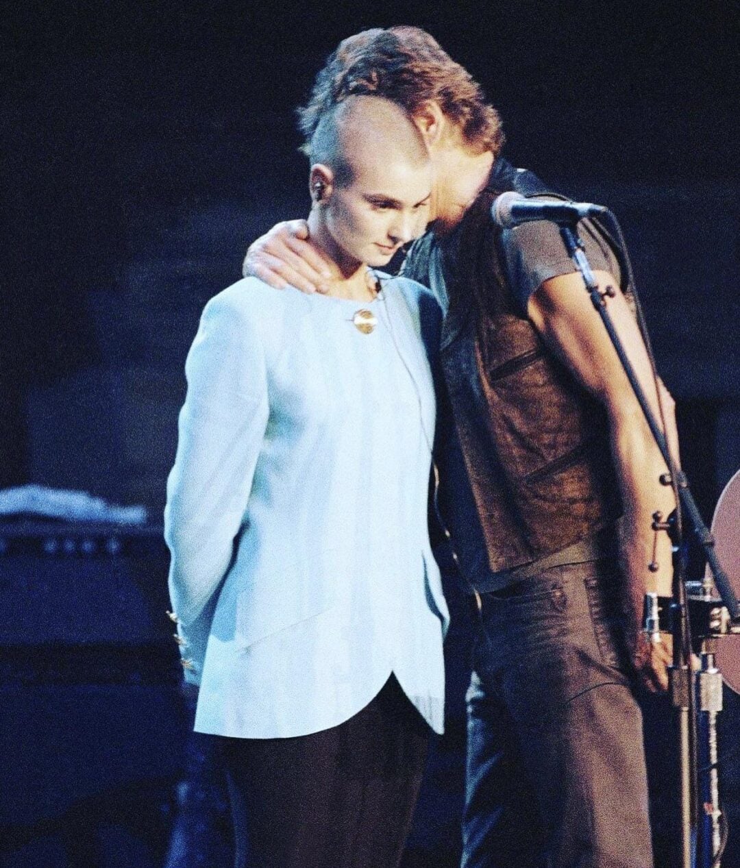 A woman with a shaved head stands on stage beside a man who has his arm around her shoulders. Both appear solemn, and they stand near a microphone, facing a dimly lit audience.