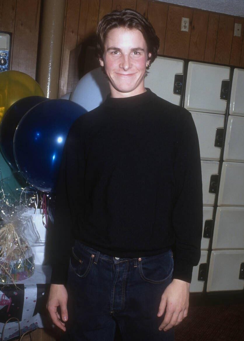 A young man with short brown hair, wearing a black long-sleeve shirt and blue jeans, stands smiling in front of lockers and colorful balloons indoors.