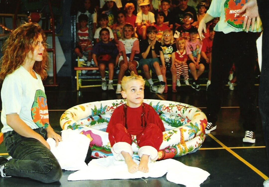 A young child in red clothes sits in a small inflatable pool, surrounded by towels, with a woman and another adult nearby. An audience of children watches in the background.