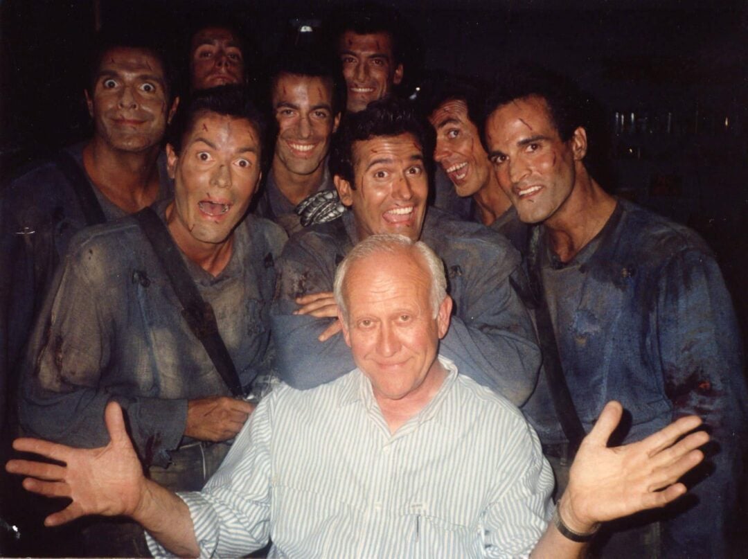 A group of seven men with dirty faces and clothes smile widely behind an older man in a striped shirt who is sitting and smiling with his arms outstretched. The group appears happy and energetic.