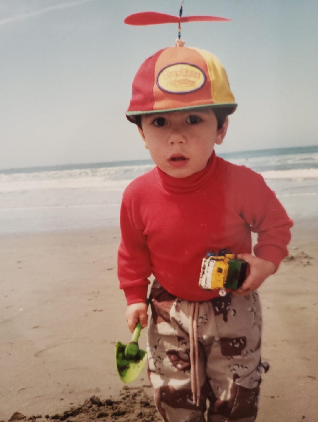 A young child in a red shirt, patterned pants, and a colorful propeller hat stands on a sandy beach holding a green shovel and toy trucks, with the ocean and sky in the background.