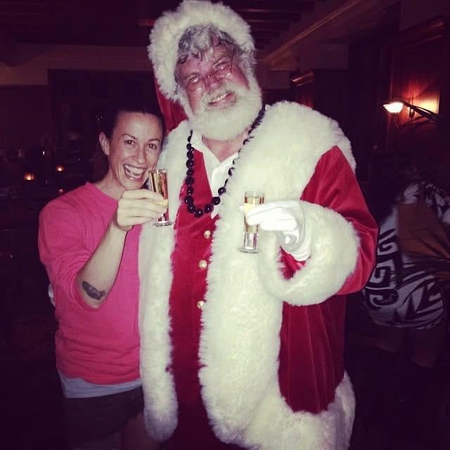 A smiling person in a pink shirt stands next to a man dressed as Santa Claus. Both are holding up shot glasses and appear to be celebrating together in a warmly lit indoor setting.