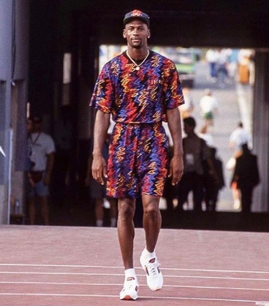 A tall man wearing a colorful, matching t-shirt and shorts set with a zigzag pattern, a cap, and white sneakers walks confidently on a track, with people and a blurred outdoor background behind him.