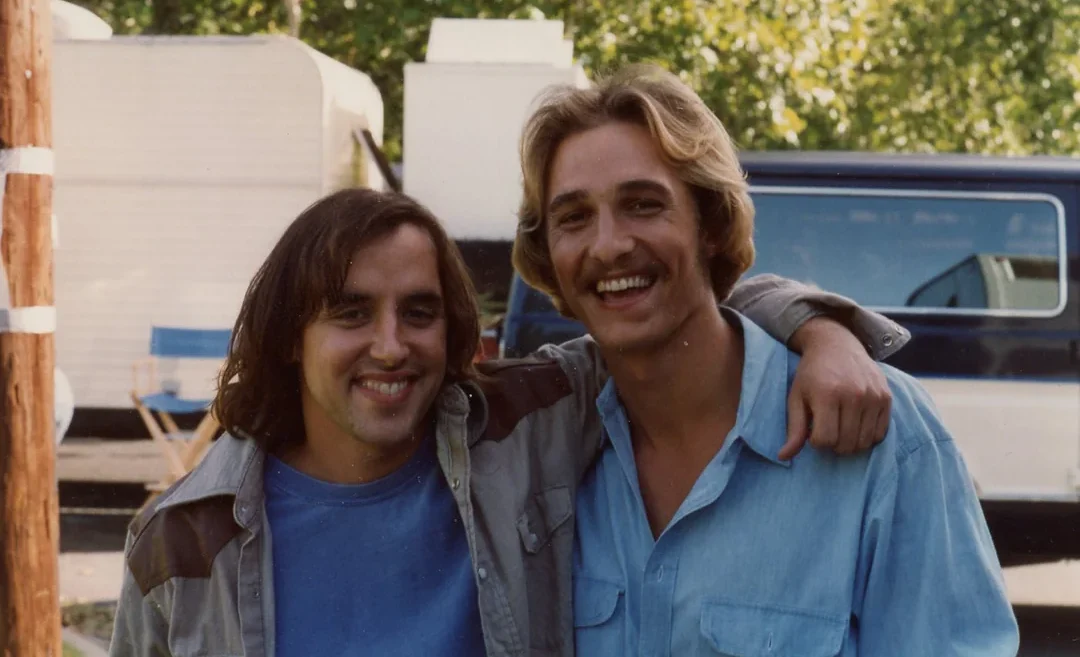 Two young men stand outdoors smiling at the camera. One has long brown hair and wears a blue shirt with a jacket, while the other has short blond hair, a mustache, and wears a light blue button-up shirt with his arm around the first man.
