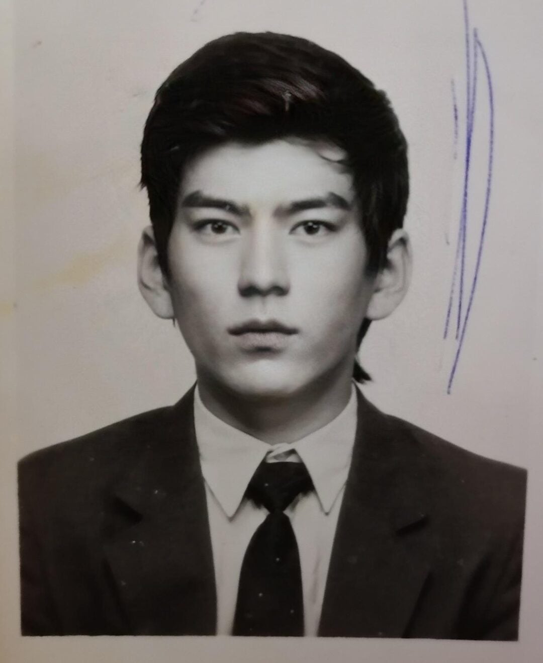 Black and white portrait of a young man with short dark hair, wearing a suit and tie, looking straight at the camera. There are blue ink marks and some discoloration on the photo's surface.