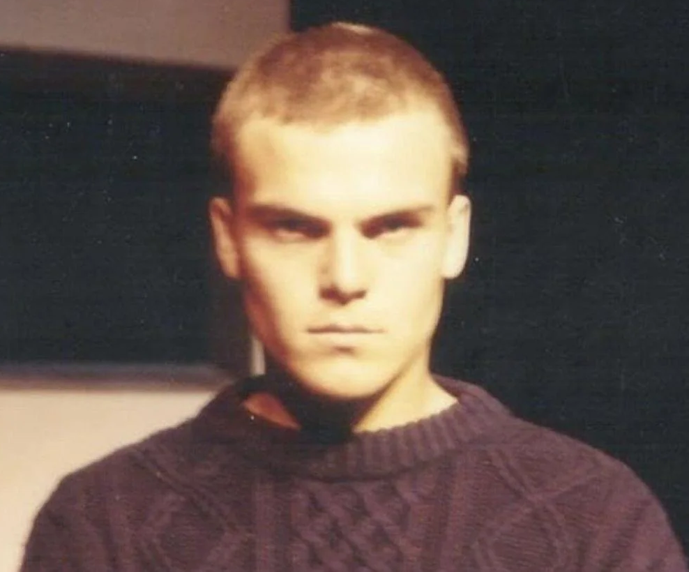 A young man with short hair, wearing a dark knit sweater, stands indoors against a blurred dark background, looking seriously at the camera.