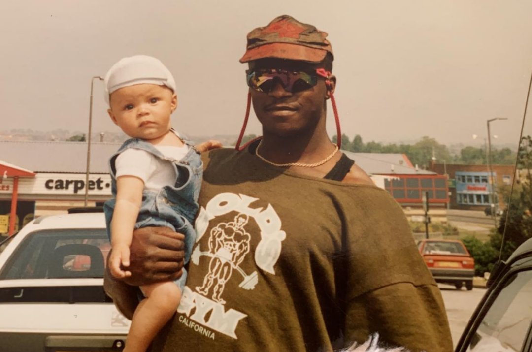 A man wearing sunglasses, a red cap, and a green t-shirt holds a baby in denim overalls and a white hat. They are standing in a parking lot with cars and store signs visible in the background.