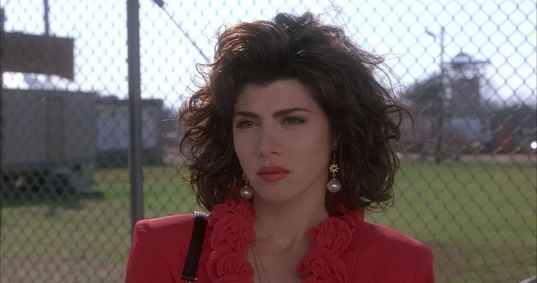 A woman with voluminous dark hair and bold red lipstick wears a bright red jacket with ruffles and large pearl earrings, standing outdoors in front of a chain-link fence and a grassy field.