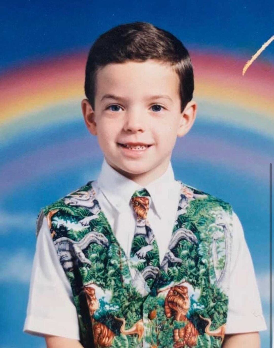 A young boy smiles in a studio portrait, wearing a white shirt and a colorful dinosaur-themed vest and tie. A vivid rainbow arches across the blue background behind him.
