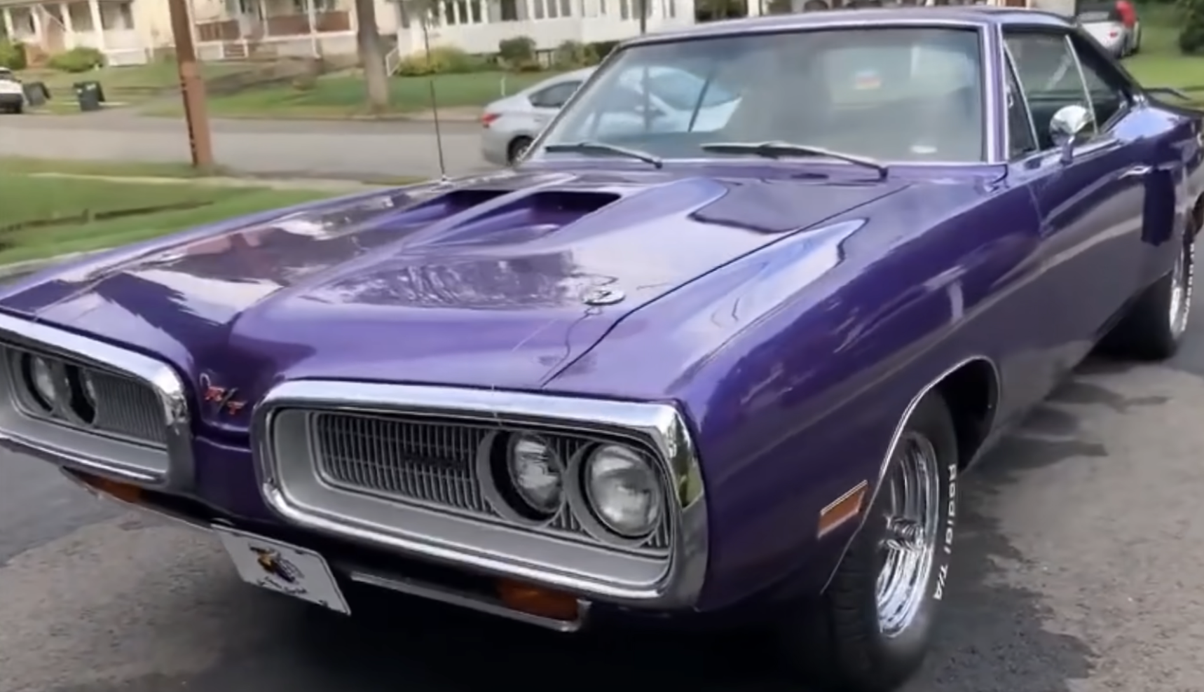 A purple classic muscle car with chrome details is parked on a suburban street, with houses and trees visible in the background.