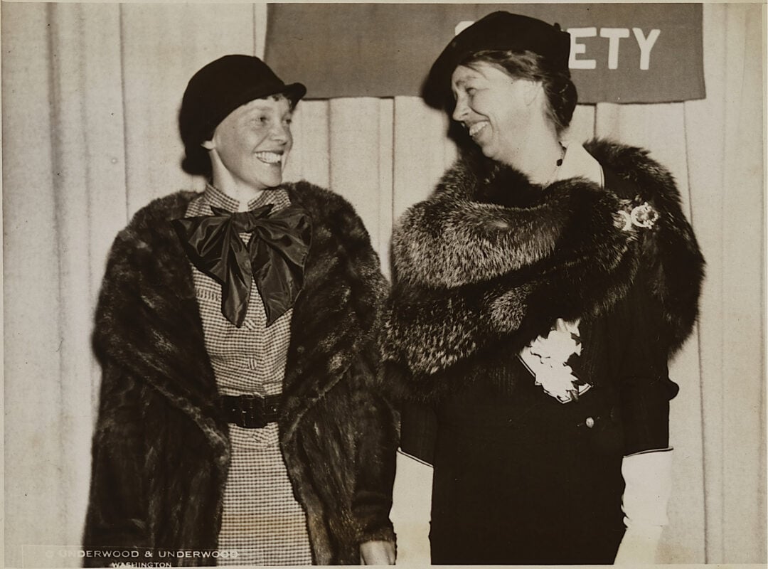 Two women wearing hats and fur stoles stand side by side, smiling at each other in front of a curtain and a partially visible sign.