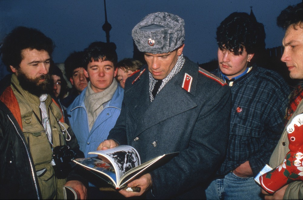 A group of men stand closely together outdoors. The central man, wearing a gray Soviet-style military coat and fur hat, looks down at an open book while others watch attentively. The sky appears dark or overcast.