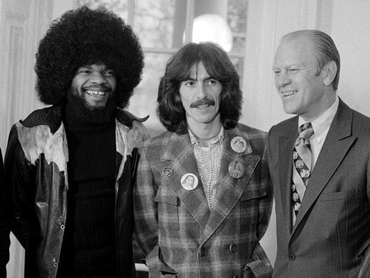 Three men stand together smiling. The man on the left has a large afro and wears a leather jacket. The man in the center has long hair and a mustache, wearing a plaid jacket with buttons. The man on the right wears a suit and tie.