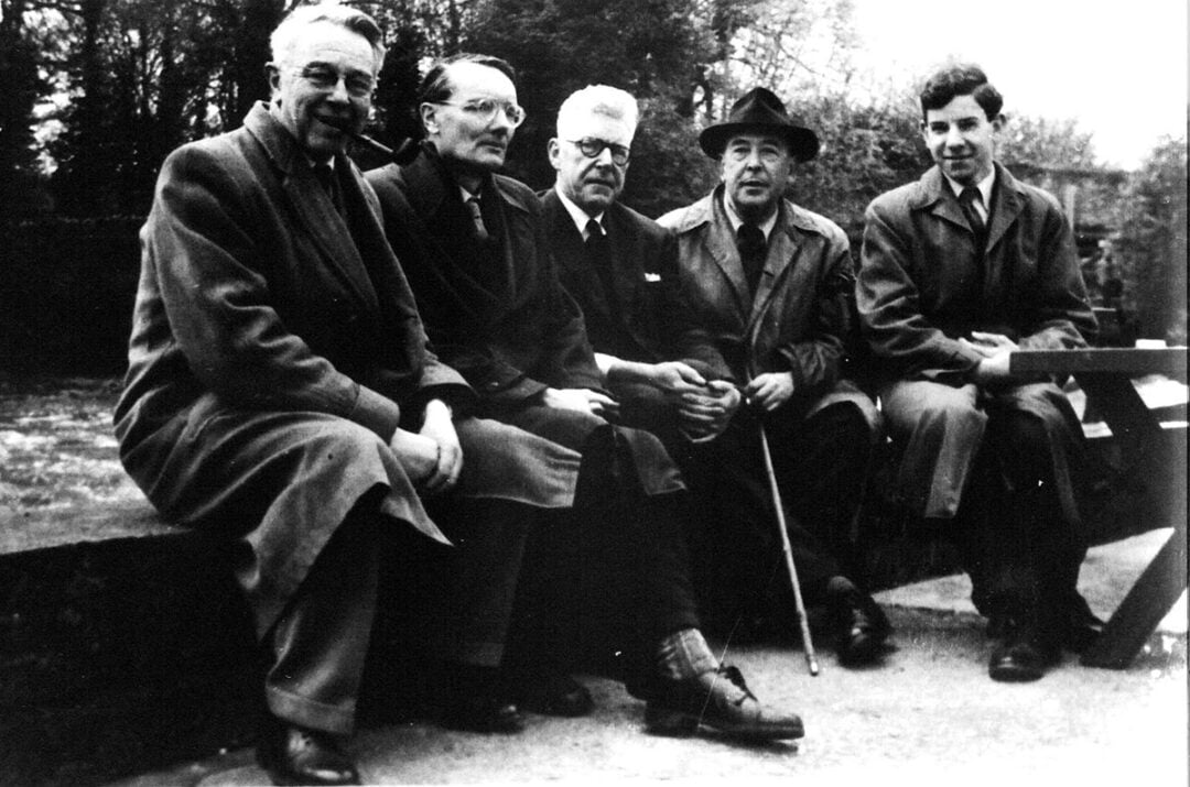 Five men wearing coats sit outdoors on a low wall and bench, facing the camera. One holds a cane. The setting appears to be a park or garden with trees and foliage in the background.