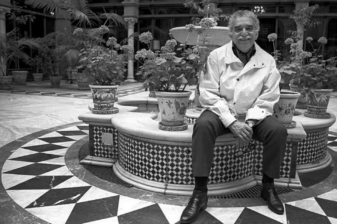 An older man in a light jacket sits smiling on the edge of a tiled fountain surrounded by potted plants in a courtyard with patterned floors and lush greenery.