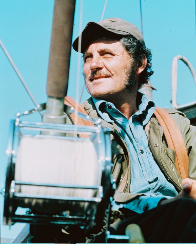 A man with a mustache wearing a cap, jacket, and denim shirt smiles while sitting on a boat, holding a fishing reel on a sunny day.