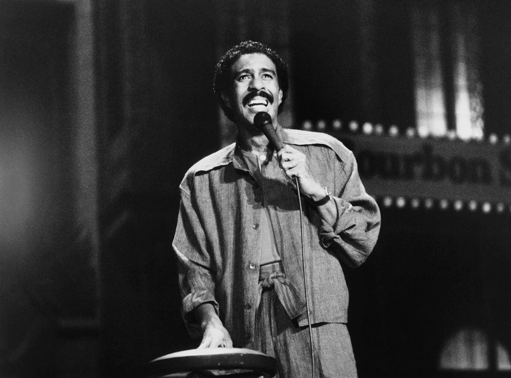 A man with a mustache stands on stage holding a microphone, wearing a loose jacket and pants, performing under a spotlight with a "Bourbon St" sign blurred in the background.