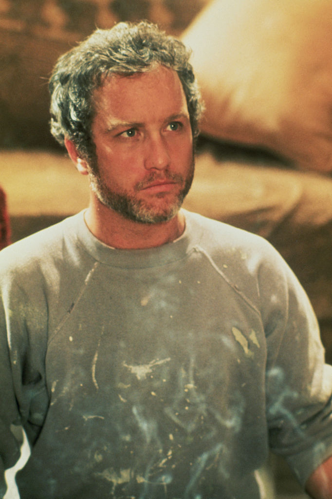 A man with curly hair and a beard wears a paint-stained sweatshirt, looking serious while standing indoors with blurred furniture in the background.