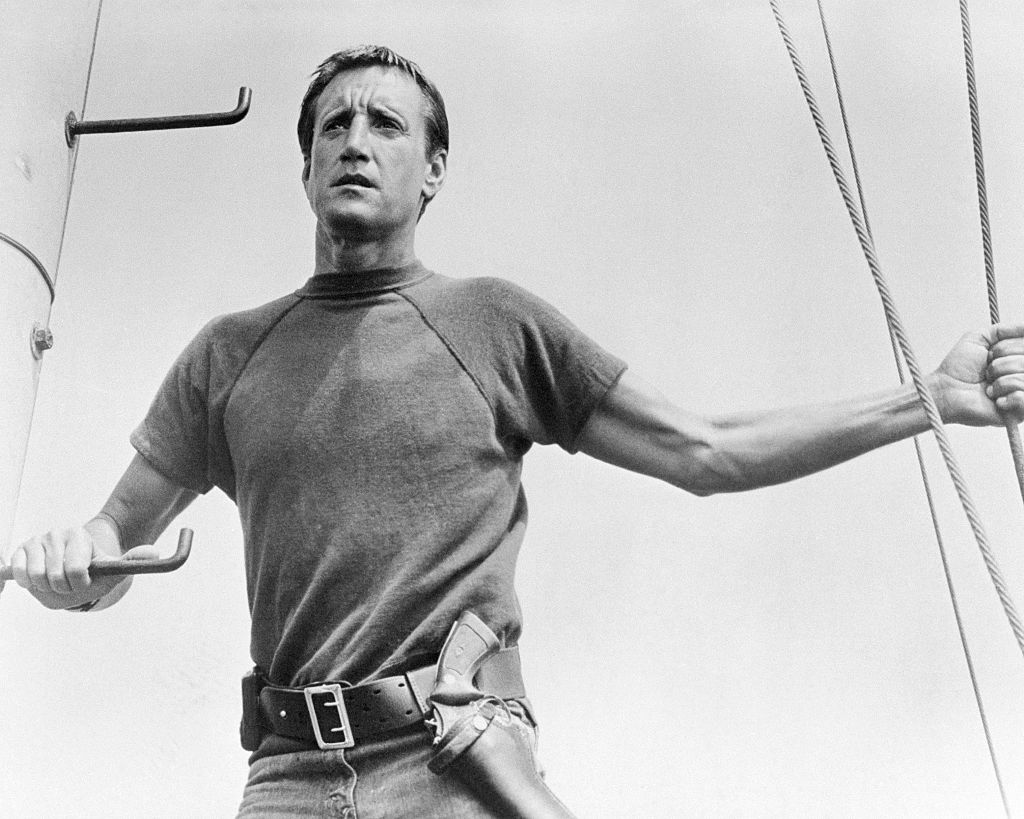 A man in a short-sleeved shirt and jeans stands on a boat, holding onto a railing with one hand and a rope with the other, looking into the distance with a serious expression. The image is in black and white.