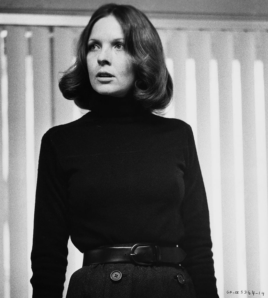A woman with shoulder-length hair, wearing a black turtleneck sweater and a belted skirt, stands in front of vertical blinds, looking slightly to the side. The photo is in black and white.