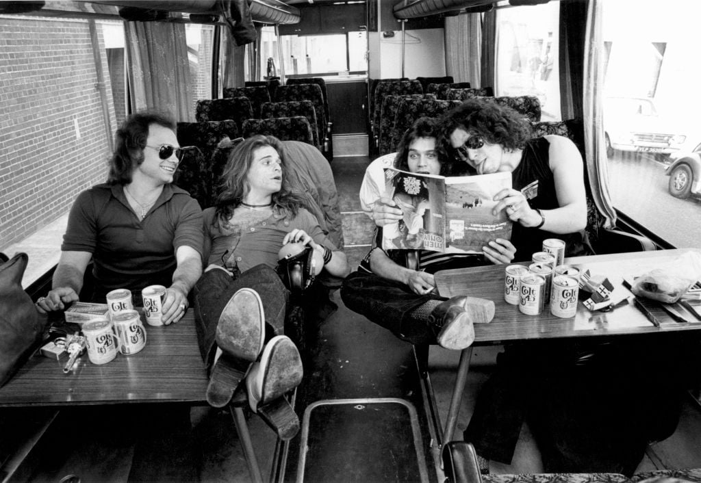 Four men relax on a tour bus, sitting around tables with beer cans. Two sit with feet up, one holds a camera, and another reads a magazine to the group. They appear to be enjoying themselves in a casual setting.