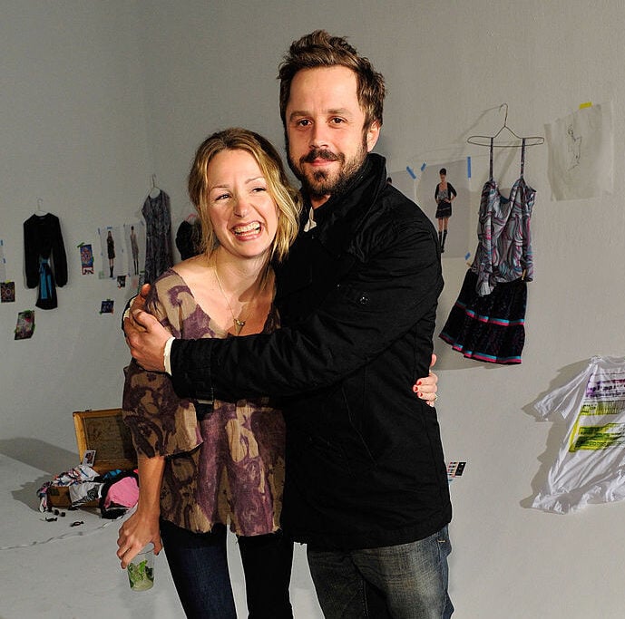 A smiling woman and man stand arm-in-arm in a white room decorated with hanging clothes, photographs, and scattered items on the floor. The woman holds a drink and both appear happy and relaxed.