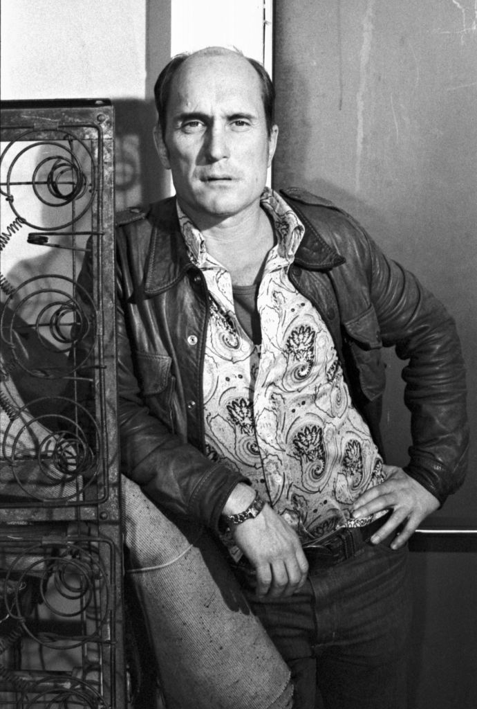 A man with a receding hairline, wearing a patterned shirt and a leather jacket, leans against a metal bed frame, looking directly at the camera with one hand on his hip. The image is in black and white.