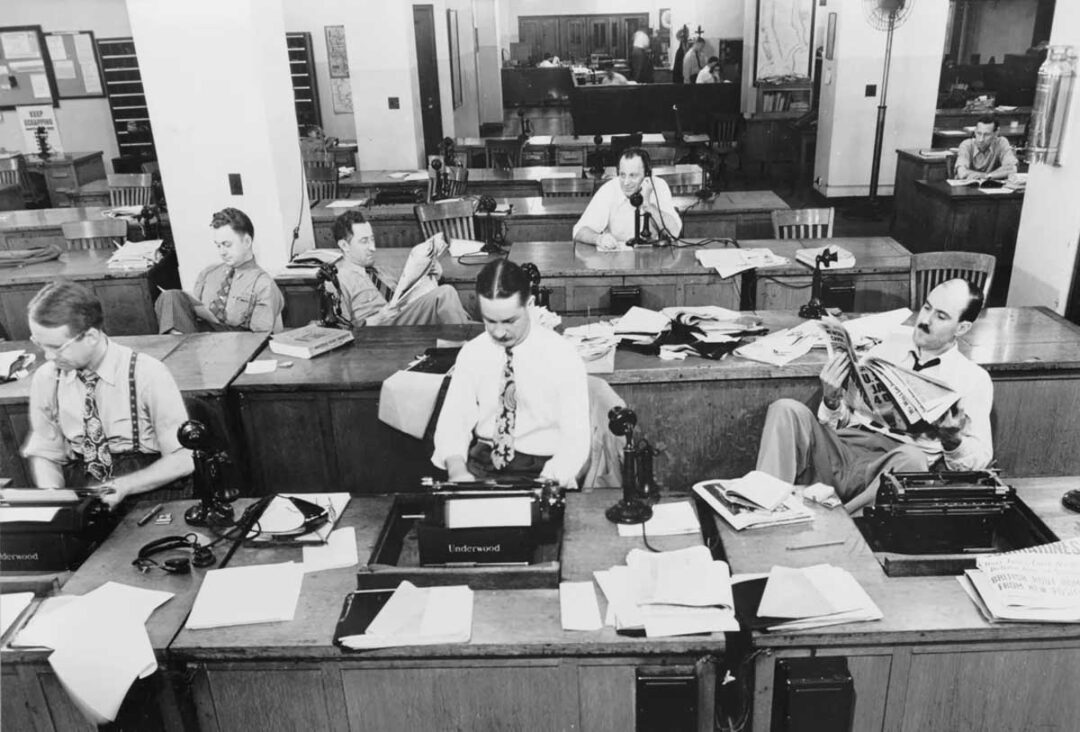 Black and white photo of a busy newsroom with men in dress shirts and ties working at desks with typewriters, papers, and phones; some are typing, others reading or talking on the phone, circa mid-20th century.