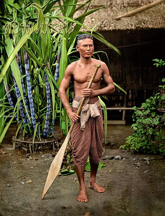 A barefoot man with short hair stands outdoors, holding a wooden paddle. He is shirtless and wears a traditional wrapped cloth around his waist. Lush green plants and a rustic building are visible in the background.