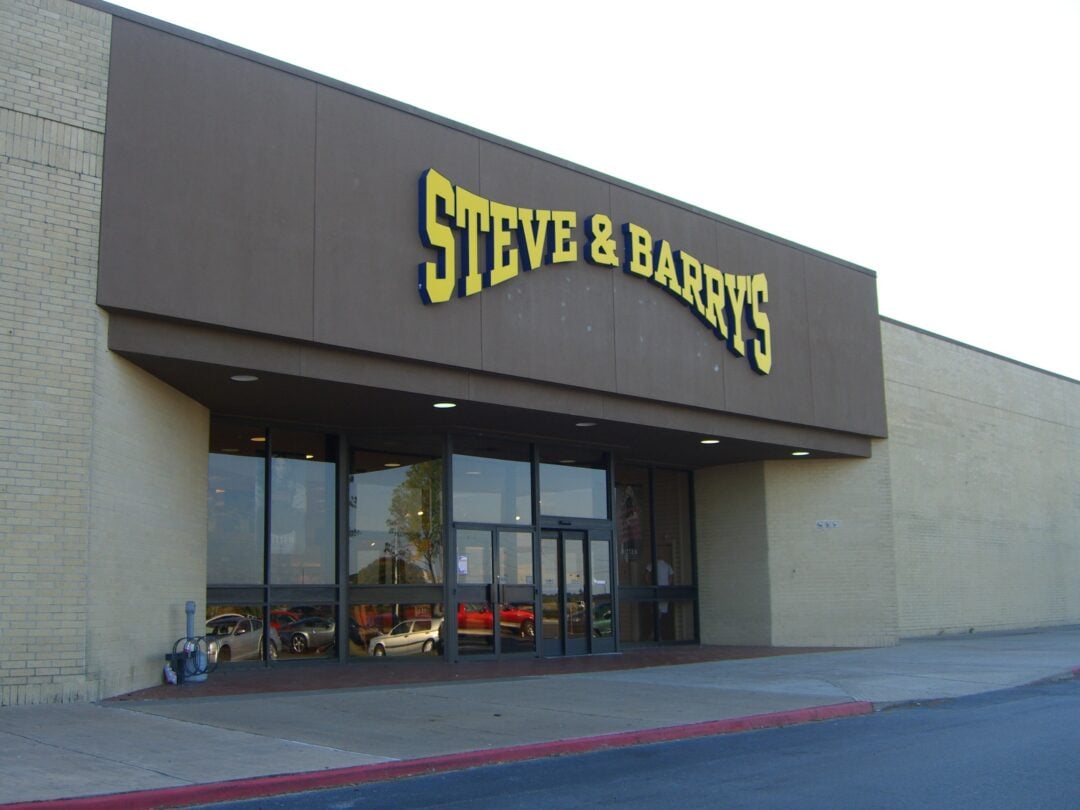 The entrance of a Steve & Barry's store with large yellow signage above glass doors, reflecting parked cars and trees outside. The building exterior is plain beige brick.