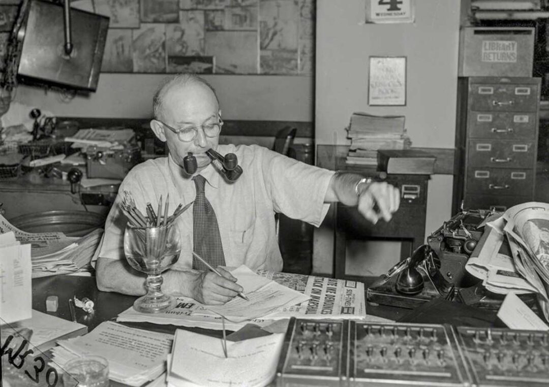 A man with glasses sits at a cluttered desk, holding two pipes in his mouth, surrounded by papers, pencils, and office equipment, while reaching for a telephone.