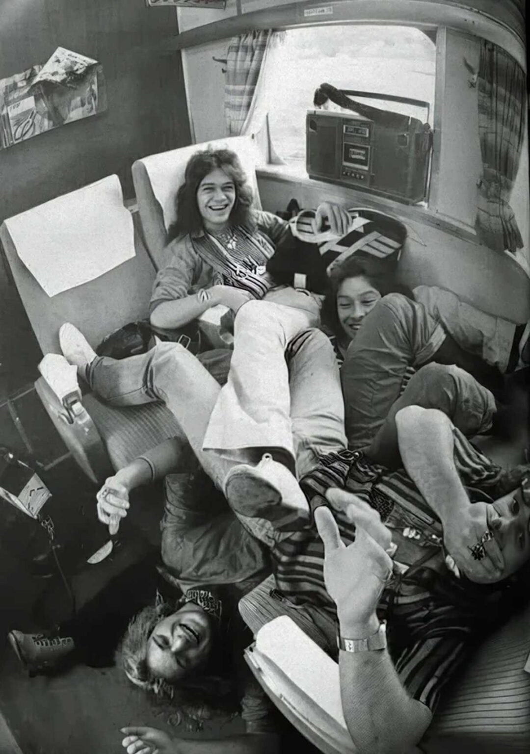 Four people are playfully piled on top of each other in a train compartment, smiling and laughing. The scene is casual and relaxed, with seats, curtains, and a window visible in the background. The image is in black and white.