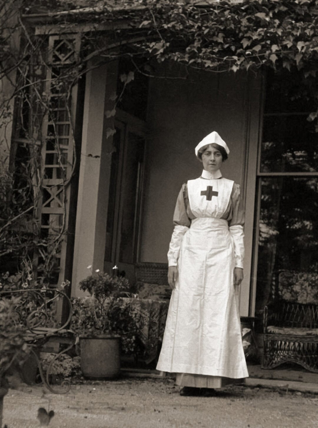 A woman in a vintage Red Cross nurse uniform stands in front of a porch, surrounded by potted plants and ivy-covered columns. The photo appears to be from the early 20th century.