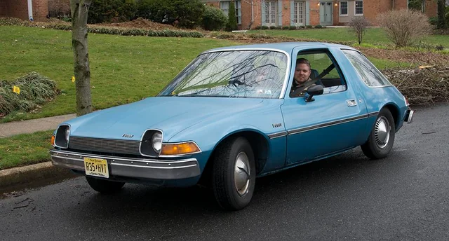 A man sits in the driver’s seat of a light blue vintage AMC Pacer parked on a suburban street, with grass and brick houses visible in the background.