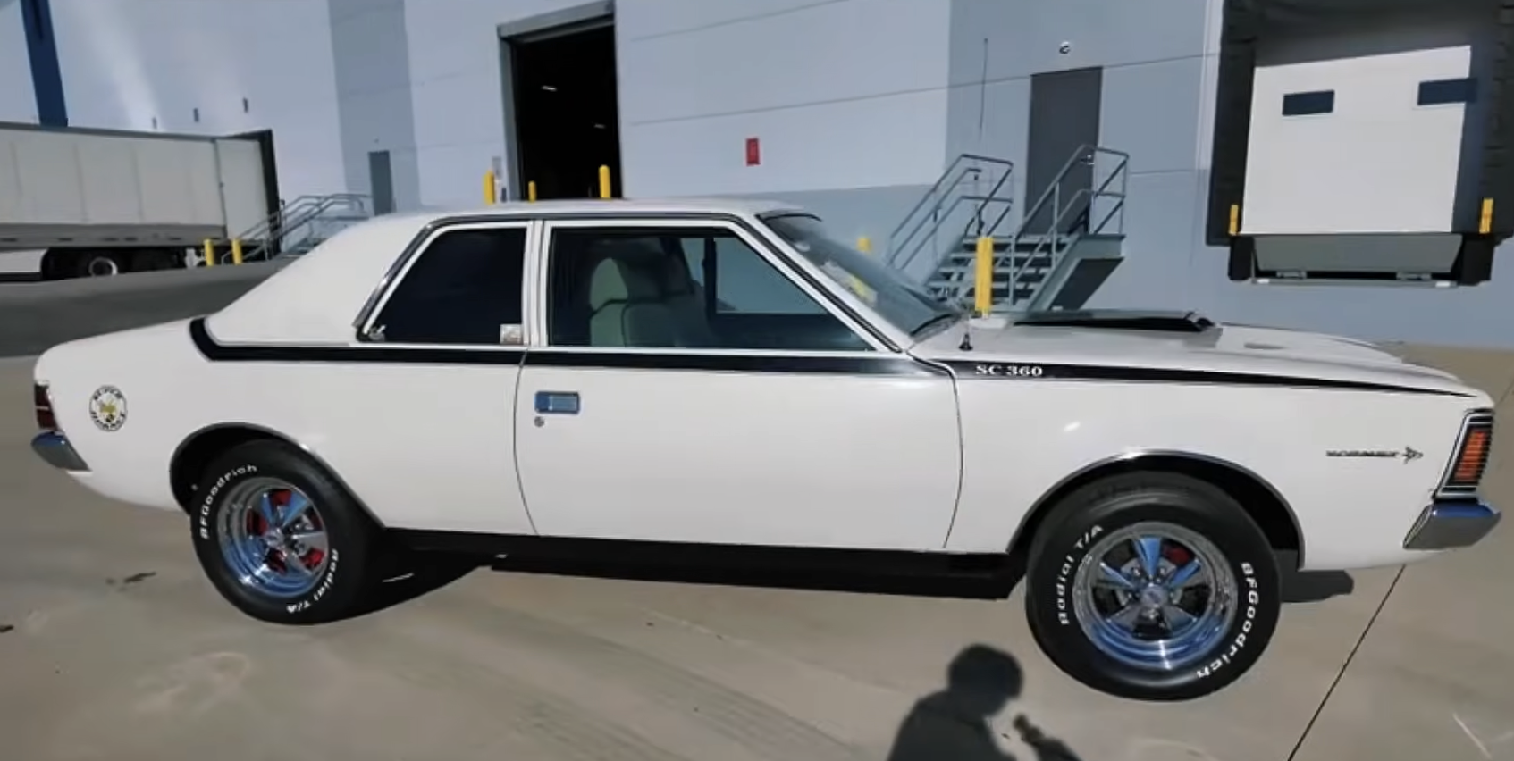 A white classic AMC car with black trim is parked in an industrial area near loading docks, with stairs and a warehouse in the background.