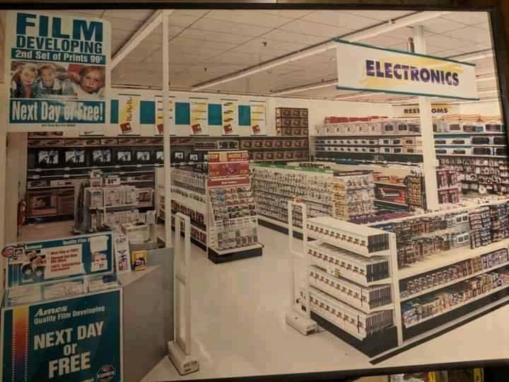 A retail store electronics section with film developing signs, shelves of tapes, DVDs, and electronics, and a sign advertising “Next Day or Free” photo prints. The area appears well-lit and organized.