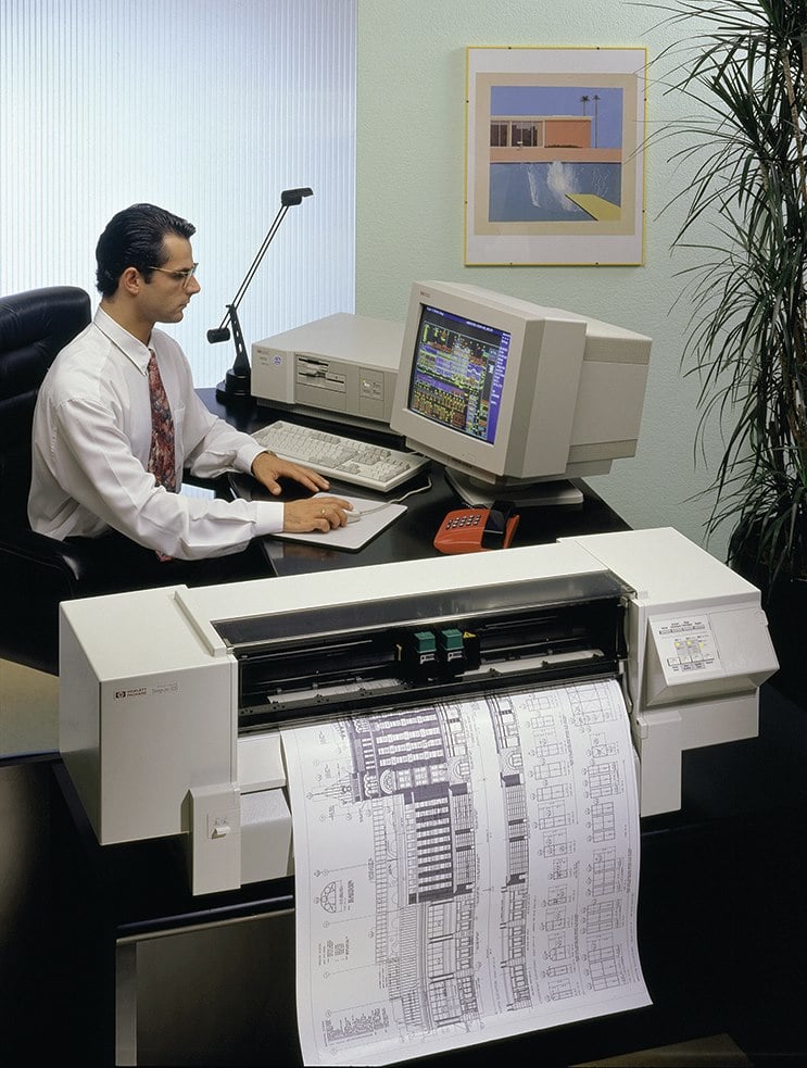 A man in an office uses a vintage desktop computer to print architectural blueprints on a large-format HP printer; a framed artwork and a plant are visible in the background.