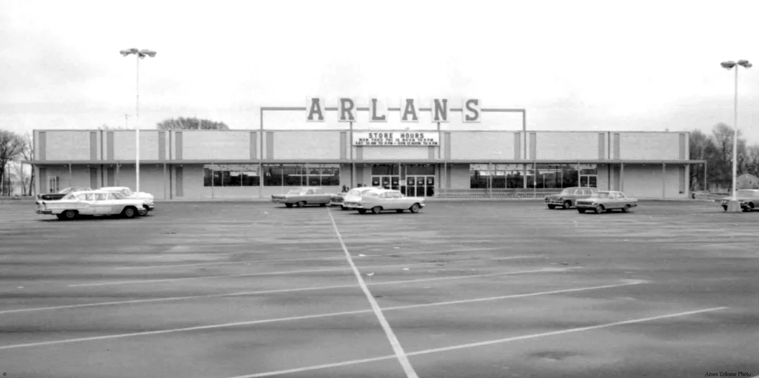 Black-and-white photo of an Arlan’s store with a large sign on the roof. The parking lot in front has a few classic 1960s cars parked, and most parking spaces are empty. Trees are visible in the background.