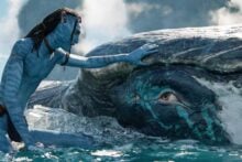 A blue-skinned humanoid character gently touches the head of a large, whale-like sea creature in the ocean, creating a moment of connection between them under a cloudy sky.
