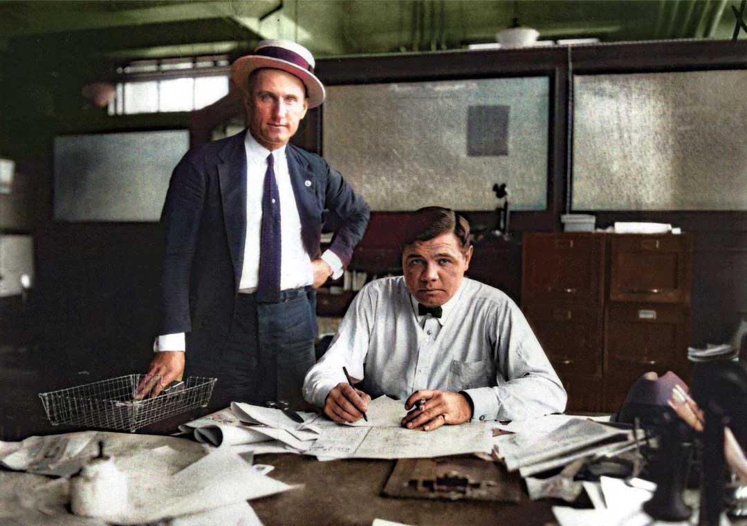 Two men in an office: one stands wearing a suit and boater hat, while the other sits at a cluttered desk, writing on papers. Both look toward the camera; office papers and a wire basket are visible.