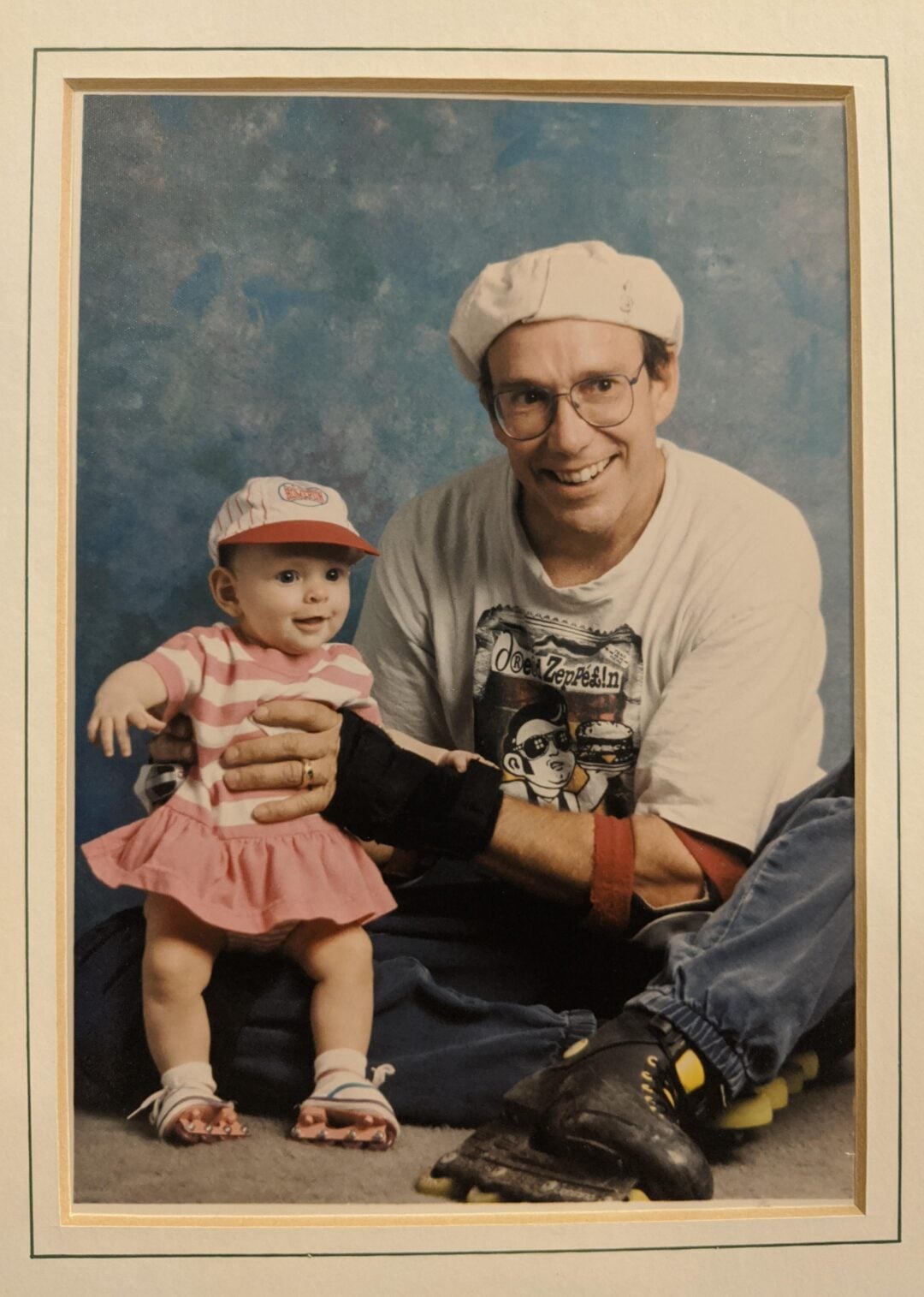 A smiling man in glasses, a white hat, and rollerblades poses with a happy baby in a pink dress and matching cap. The man holds the baby steady as they both sit against a blue portrait backdrop.