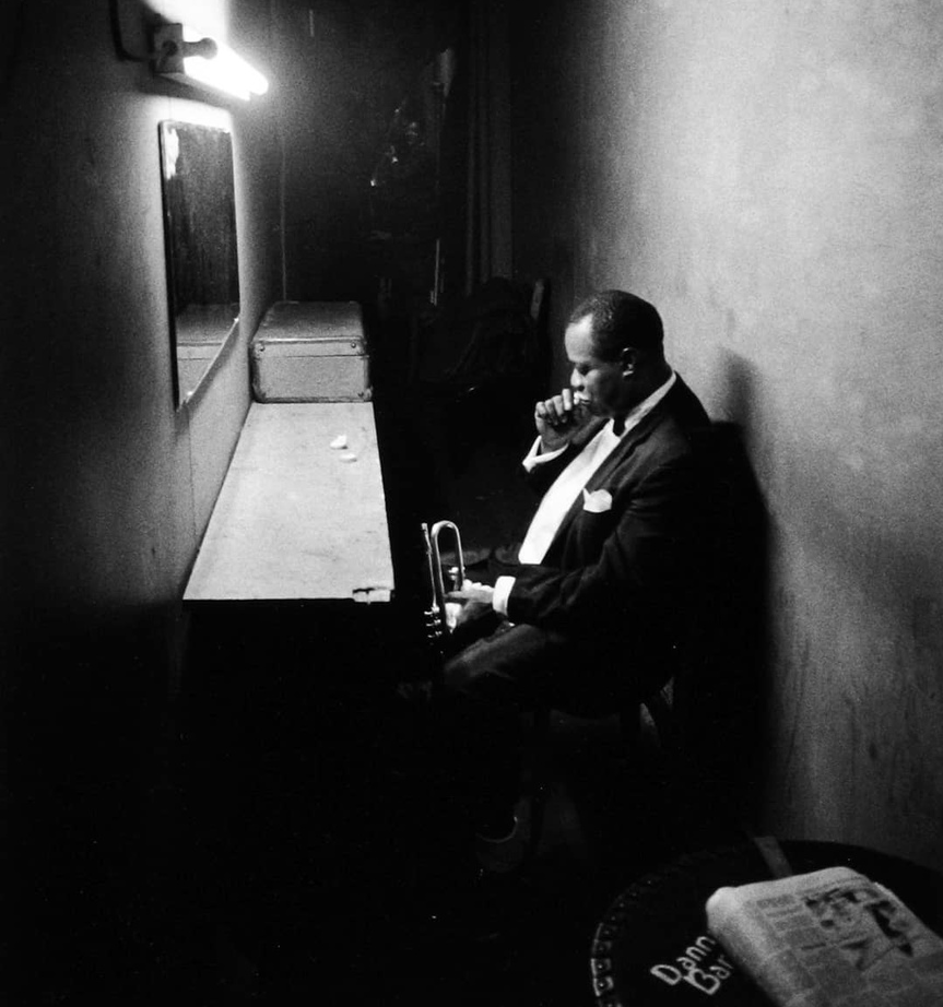 A man in a suit sits alone in a dimly lit backstage dressing room, holding a trumpet and appearing contemplative. A mirror, bench, and overhead light are visible, with a newspaper on a table in the foreground.