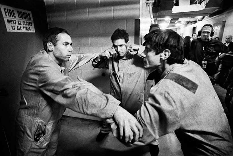 Three men in jumpsuits stand tensely in a narrow hallway, gripping each other’s arms. Their expressions are intense. Other people and a "Fire Doors Must Be Closed at All Times" sign are visible in the background.
