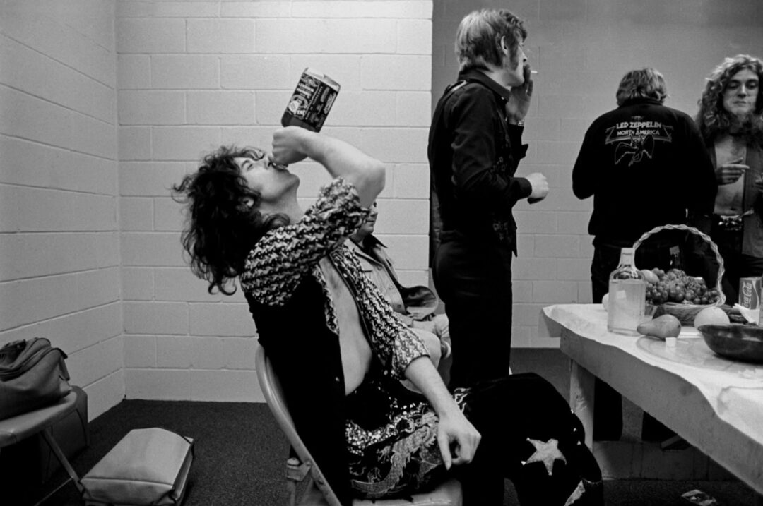 A person in star-patterned clothes tilts back a bottle to drink backstage; others stand and talk near a table with fruit and drinks. One person wears a Led Zeppelin jacket. The scene feels candid and lively.