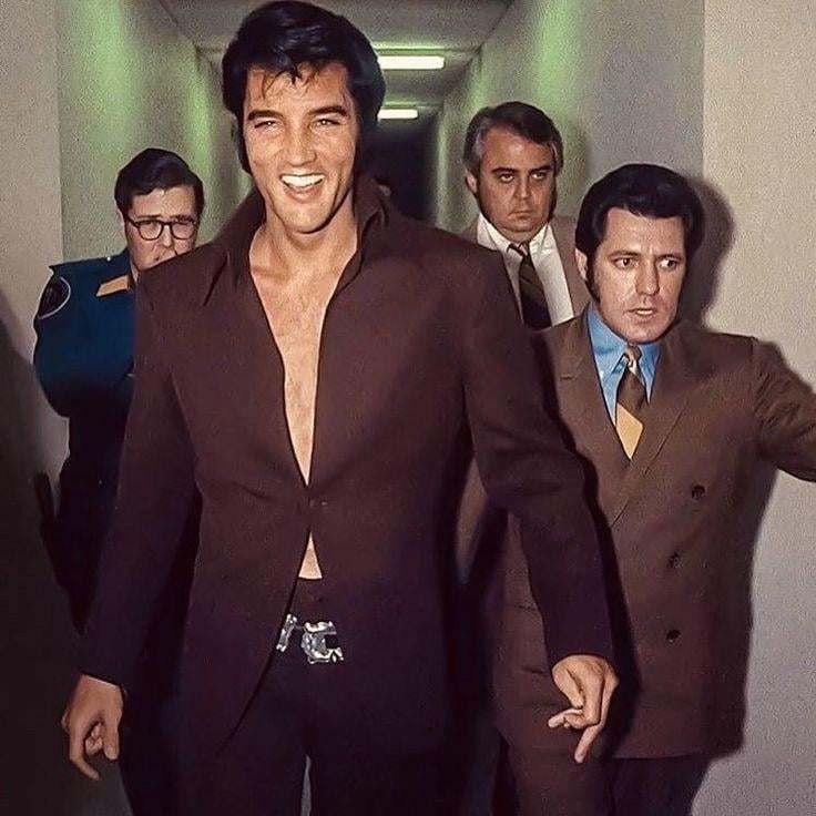 A man with dark hair and a wide smile walks down a hallway, wearing a dark brown unbuttoned jacket. He is flanked by three serious-looking men in suits, one in uniform, and bright lights illuminate the corridor.