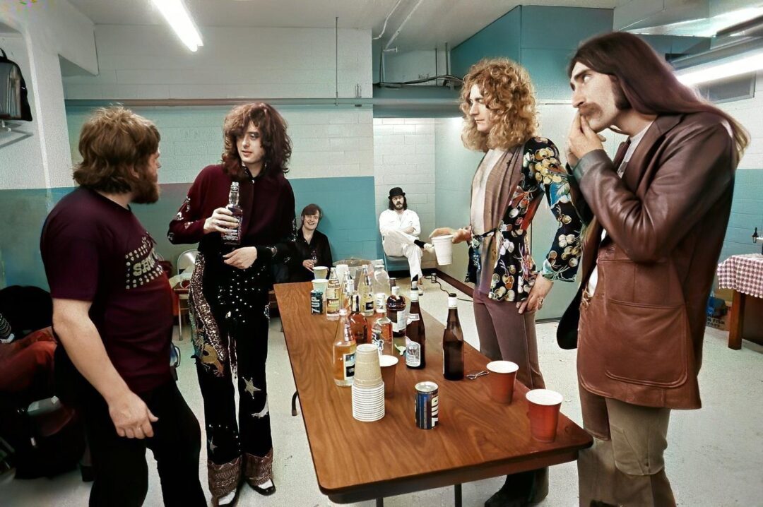 Four men stand around a table with drinks in a backstage room. One holds a bottle, another wears a floral suit, while others dress in 1970s fashion. The room has blue walls and fluorescent lighting.
